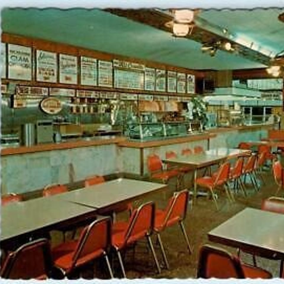Retro diner interior with red chairs, light-colored tables, and a long counter with menu boards.