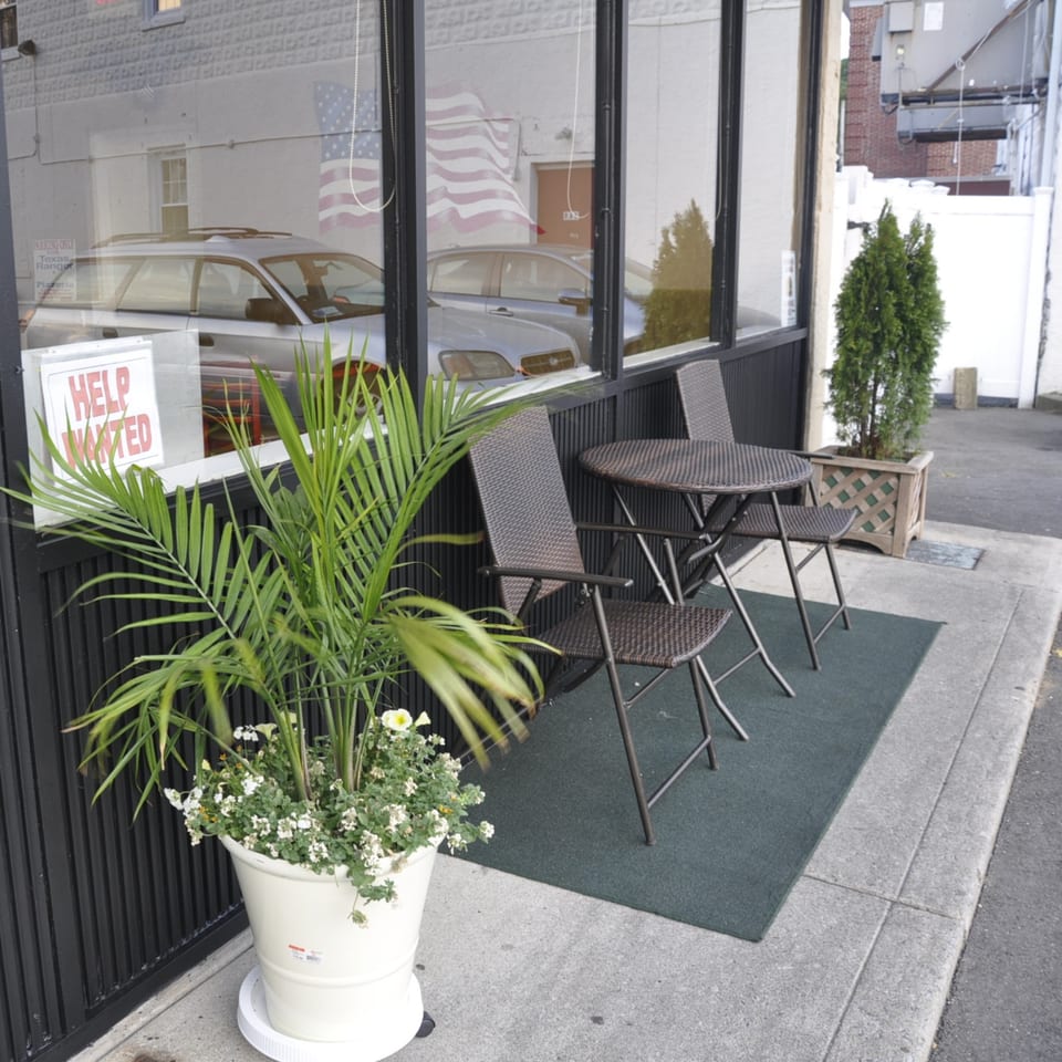 Outdoor seating area with two tables and chairs, a large potted plant, and a "Help Wanted" sign.
