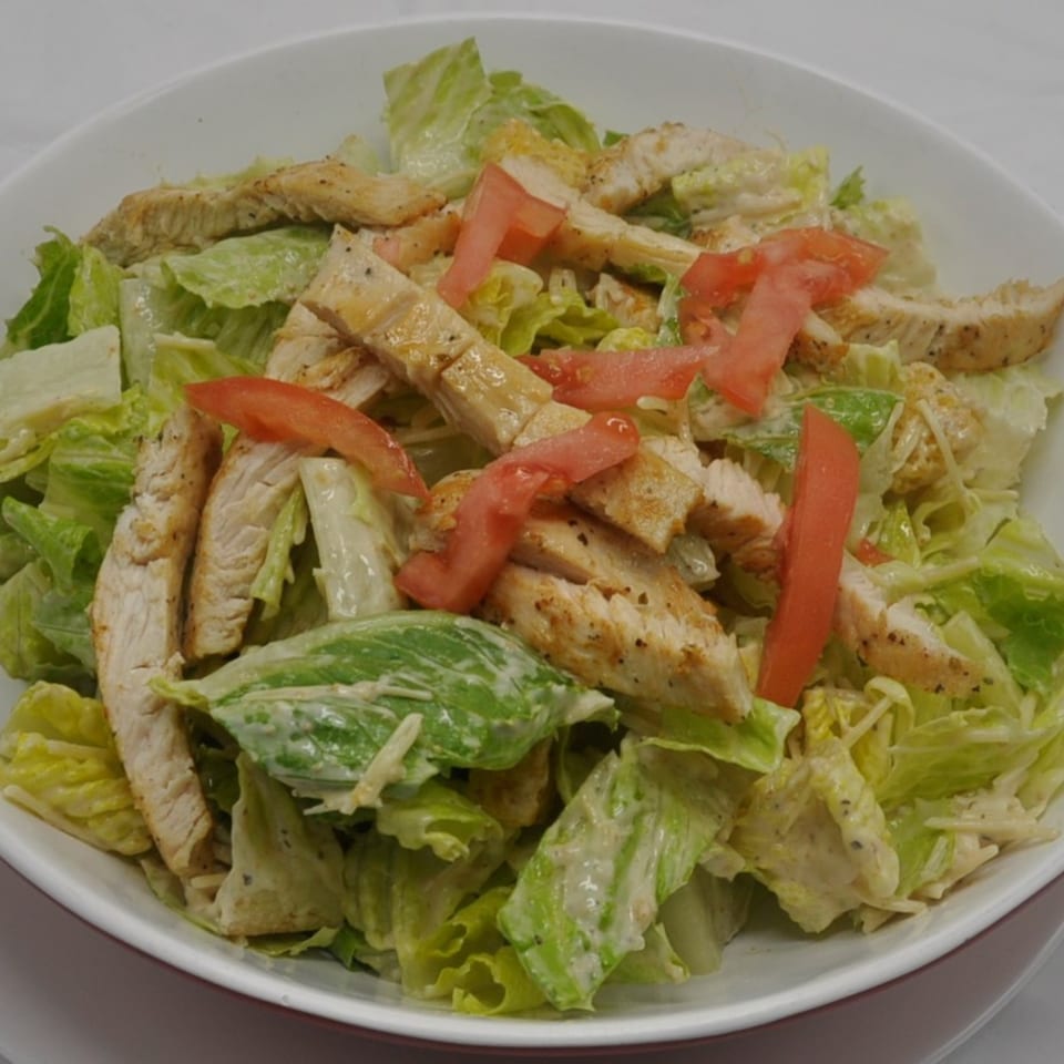 A Caesar salad with grilled chicken strips and tomato wedges in a white bowl.