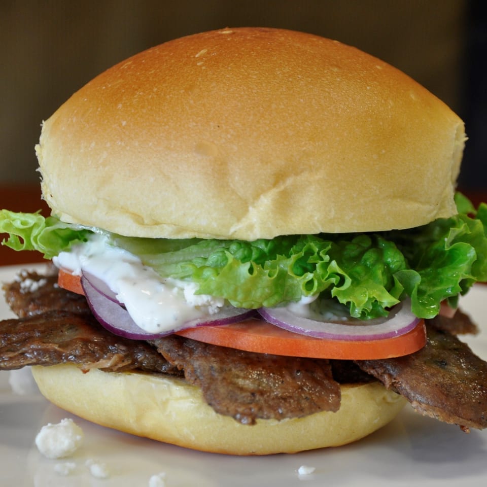 A Texas Ranger burger with sliced meat, lettuce, tomato, red onion, and white sauce on a bun.